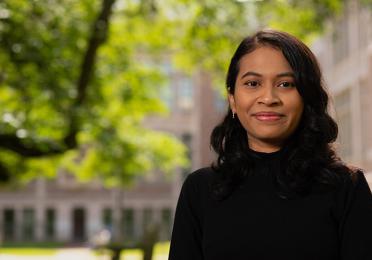 An image of Anselma Prihandita, wearing a black turtleneck and standing against a backdrop of the quad